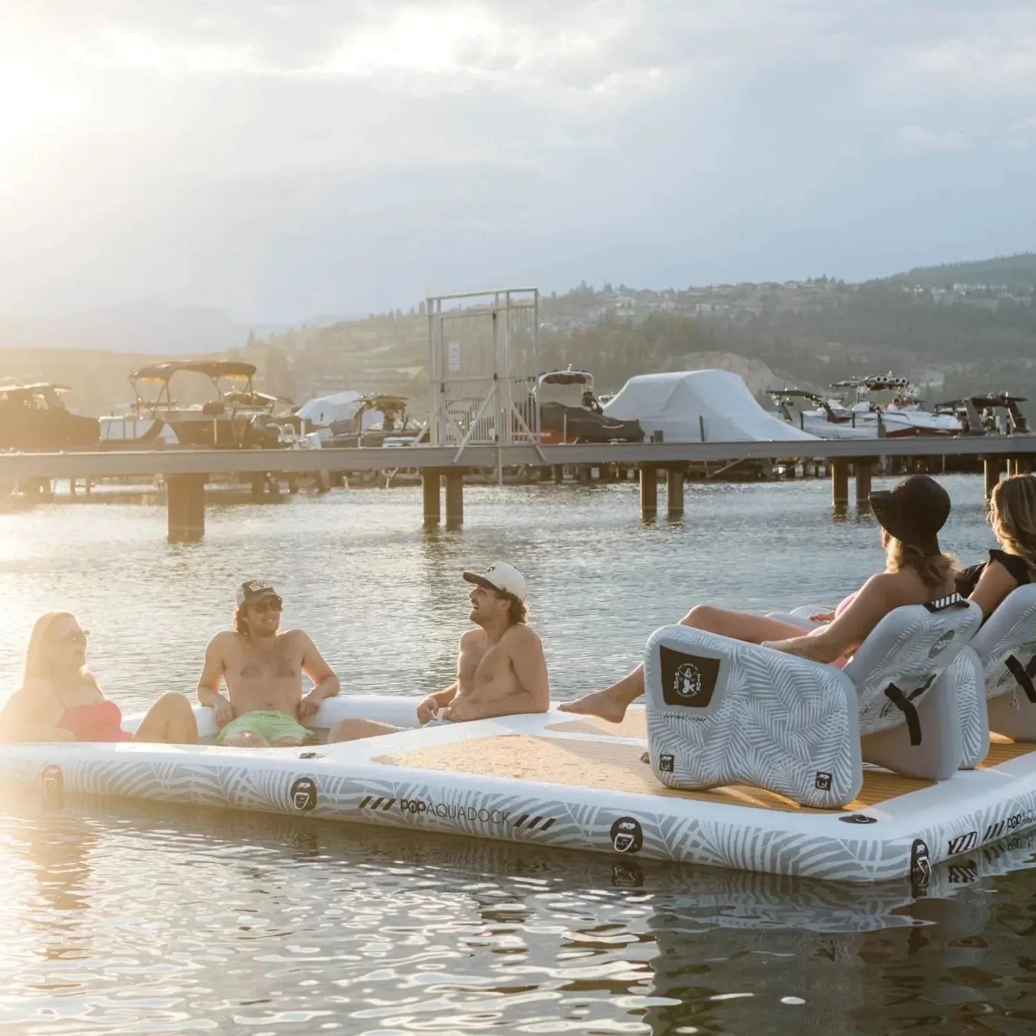 Group of friends lounging on an Aqua Dock inflatable platform with chairs, enjoying a sunny day on the water near a marina.