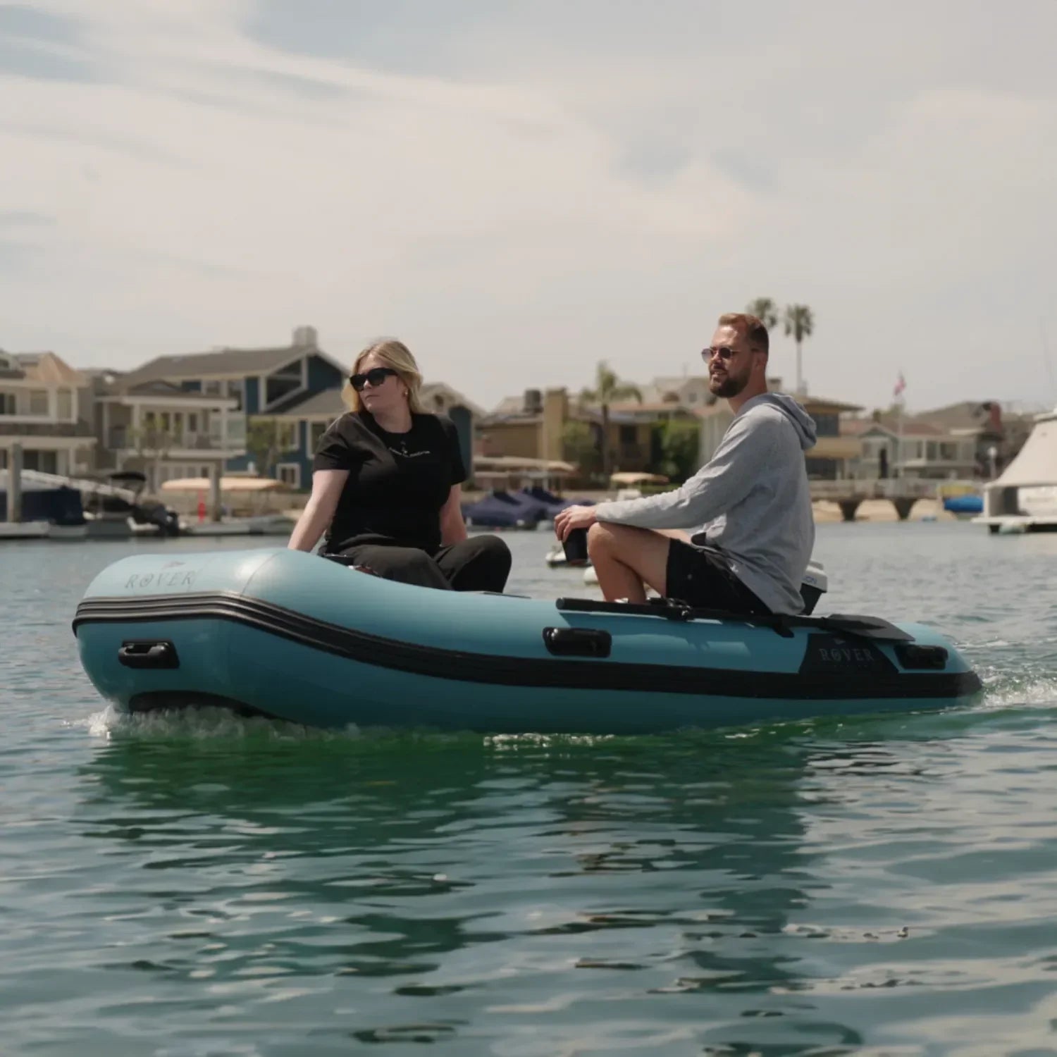 A couple relaxing and cruising on a Rover Marine Battle Boat inflatable boat, enjoying calm water with waterfront homes in the background.