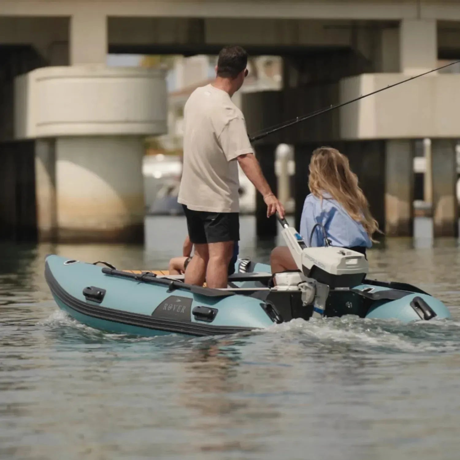 A couple fishing on a Rover Marine Battle Cat inflatable boat, powered by an ePropulsion electric outboard motor, while cruising under a bridge on calm water.