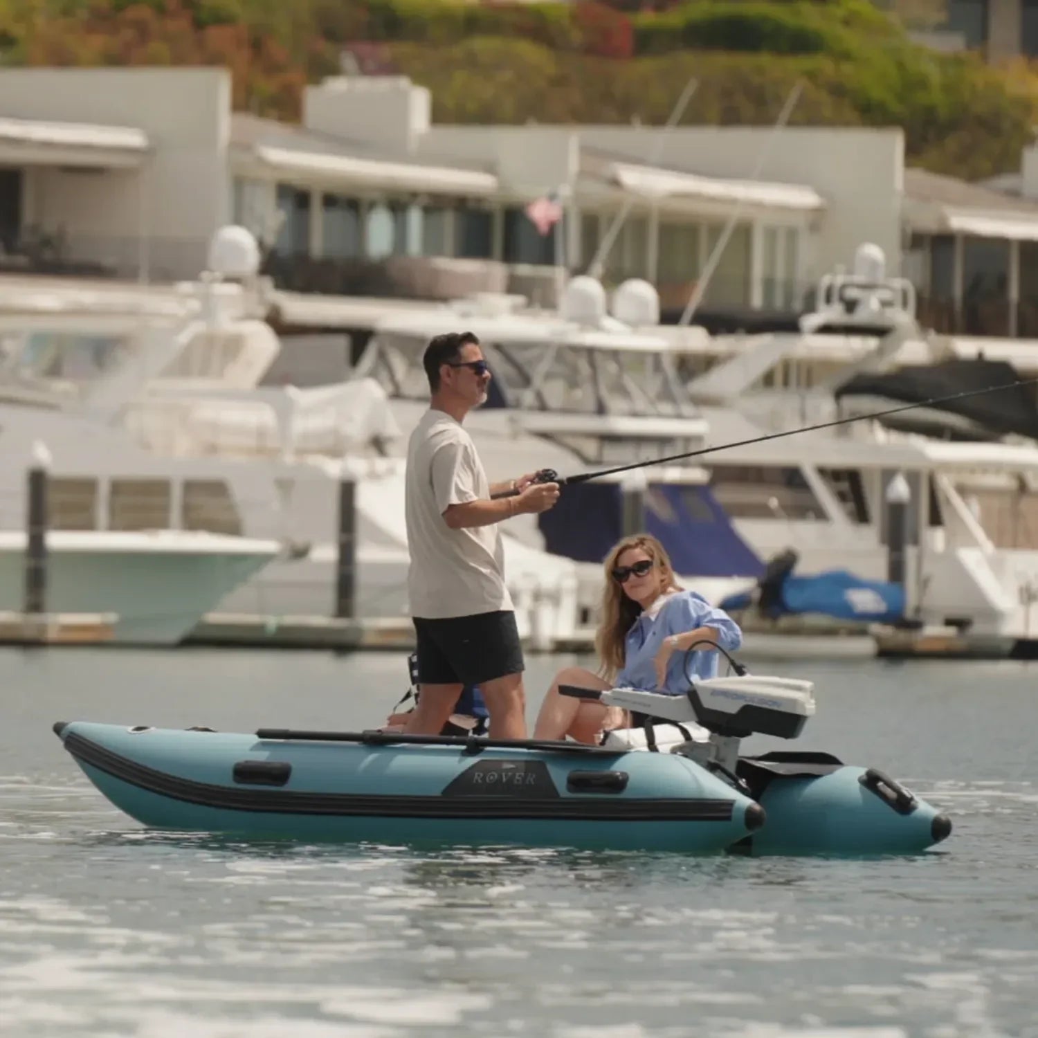 A man and woman fishing on a Rover Marine Battle Cat inflatable boat, equipped with an ePropulsion electric motor, while anchored near a marina with yachts in the background.