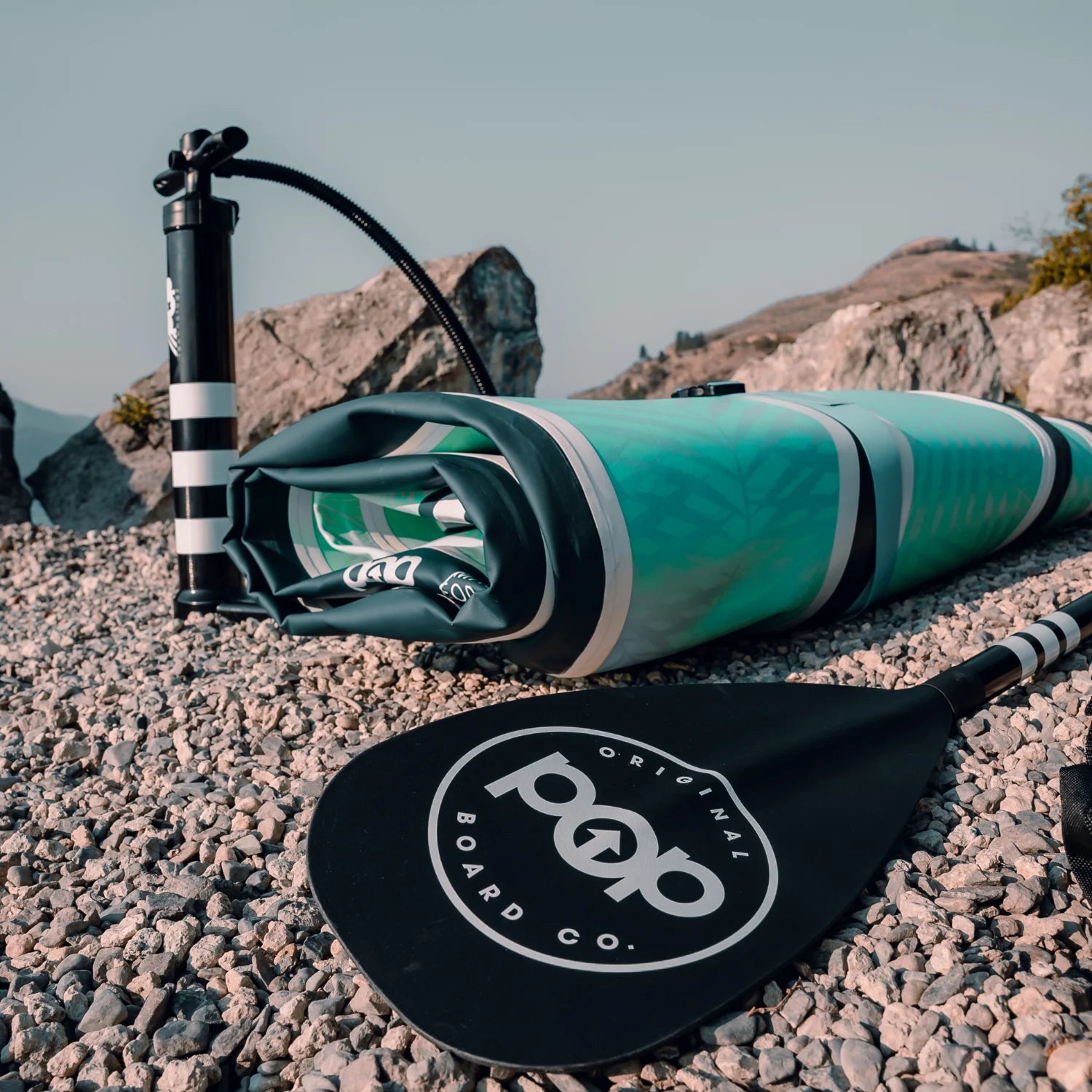 Folded POP Yacht Hopper inflatable paddleboard with hand pump and paddle on rocky shore.