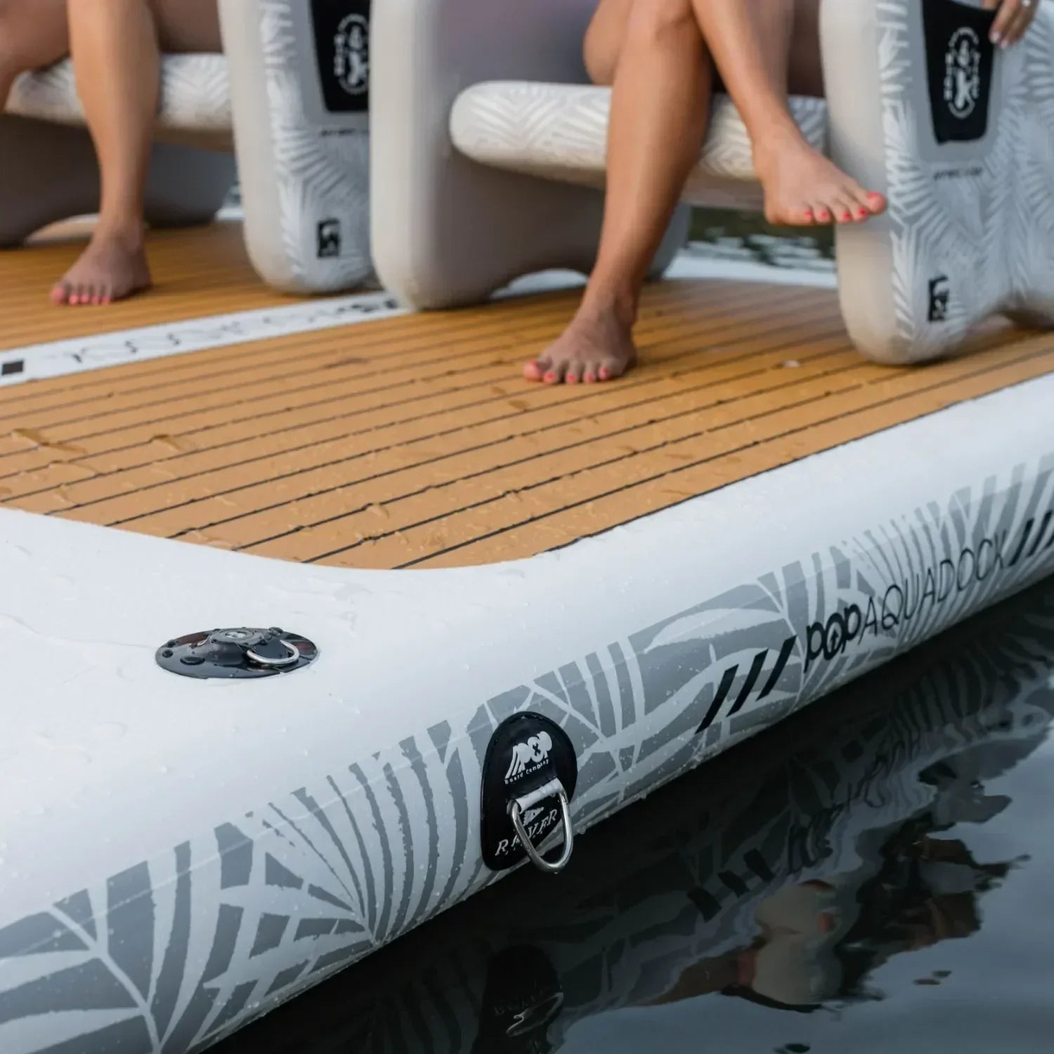 Close-up of the POP Aquadock inflatable platform showing its premium non-slip PVC traction pad and anchor points, with people sitting comfortably on inflatable chairs.