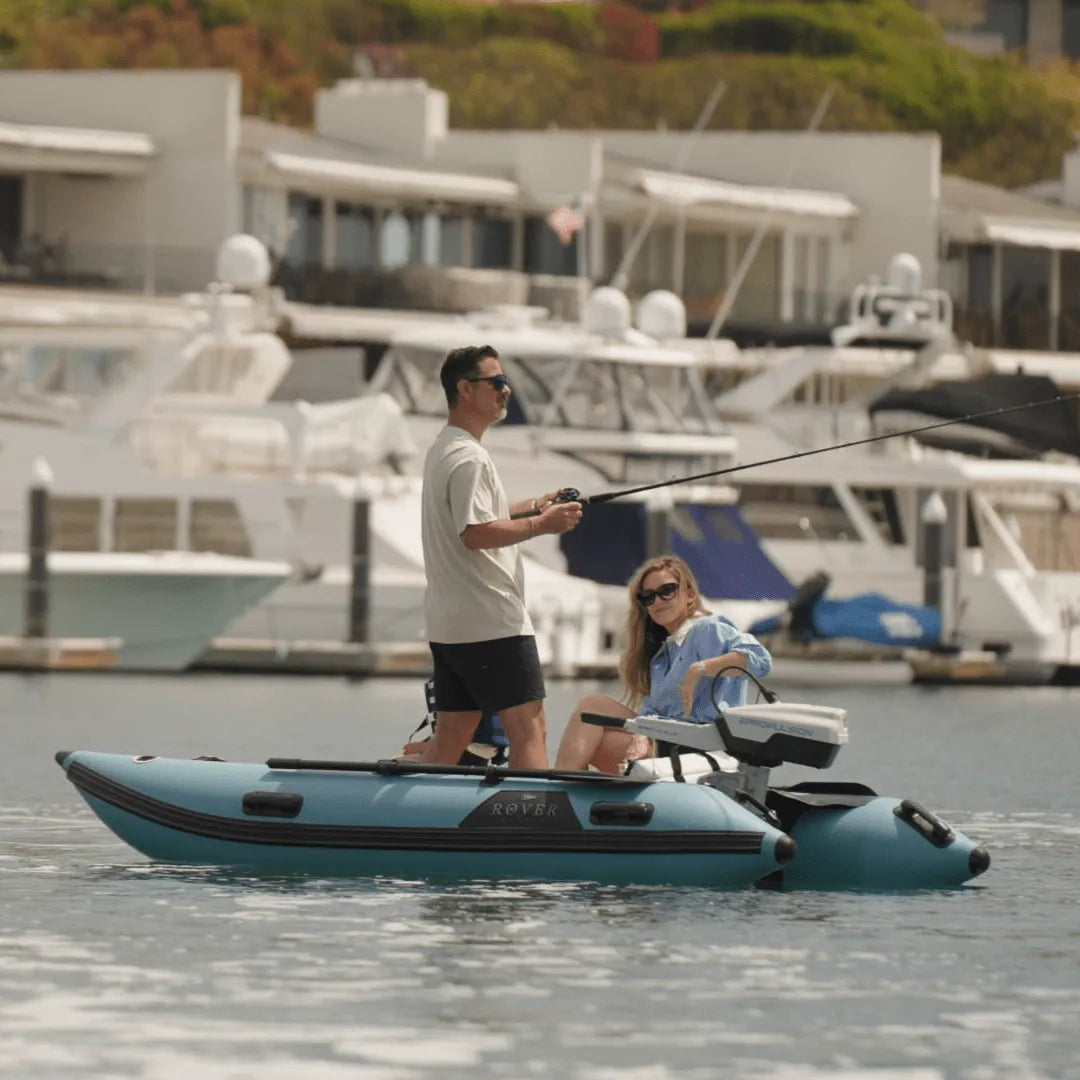 Two people on a small inflatable boat in a marina with boats and buildings in the background.