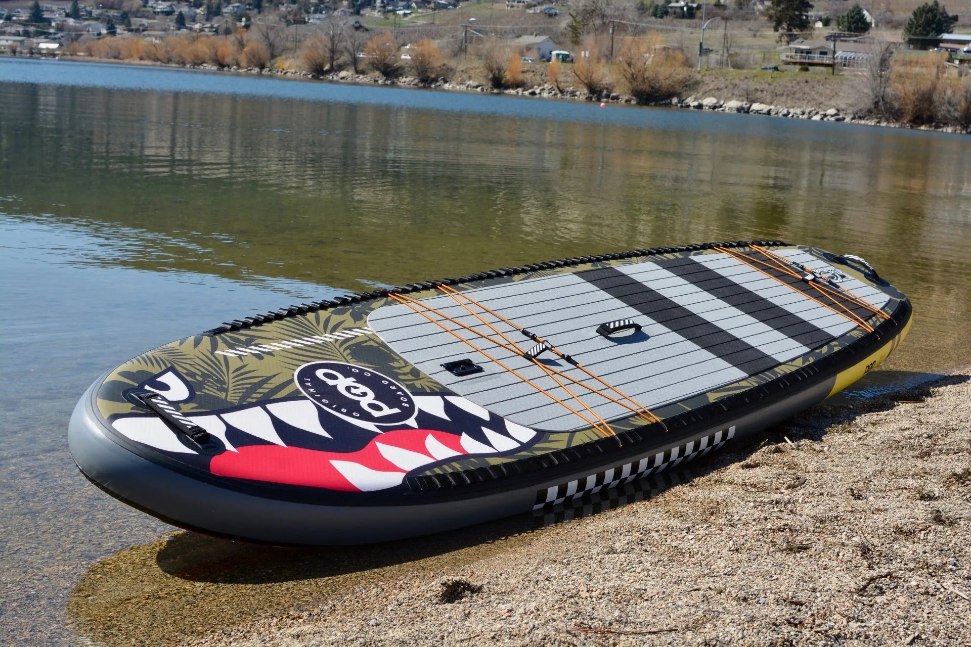 Full view of the 11'6" El Capitan Bomber inflatable paddleboard resting on a gravel shoreline, showcasing the wide 36-inch deck, shark mouth graphics, and cargo tie-down system.
