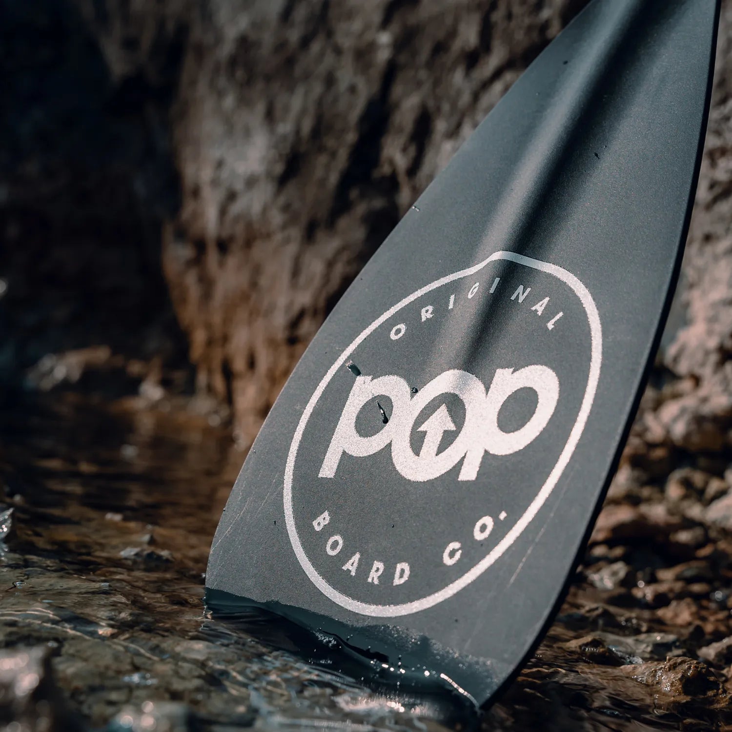 A close-up of the POP 3 pc Aluminum Collapsible Paddle's black blade, featuring the pop Original Board Co. logo, partially submerged in shallow water near rocks.