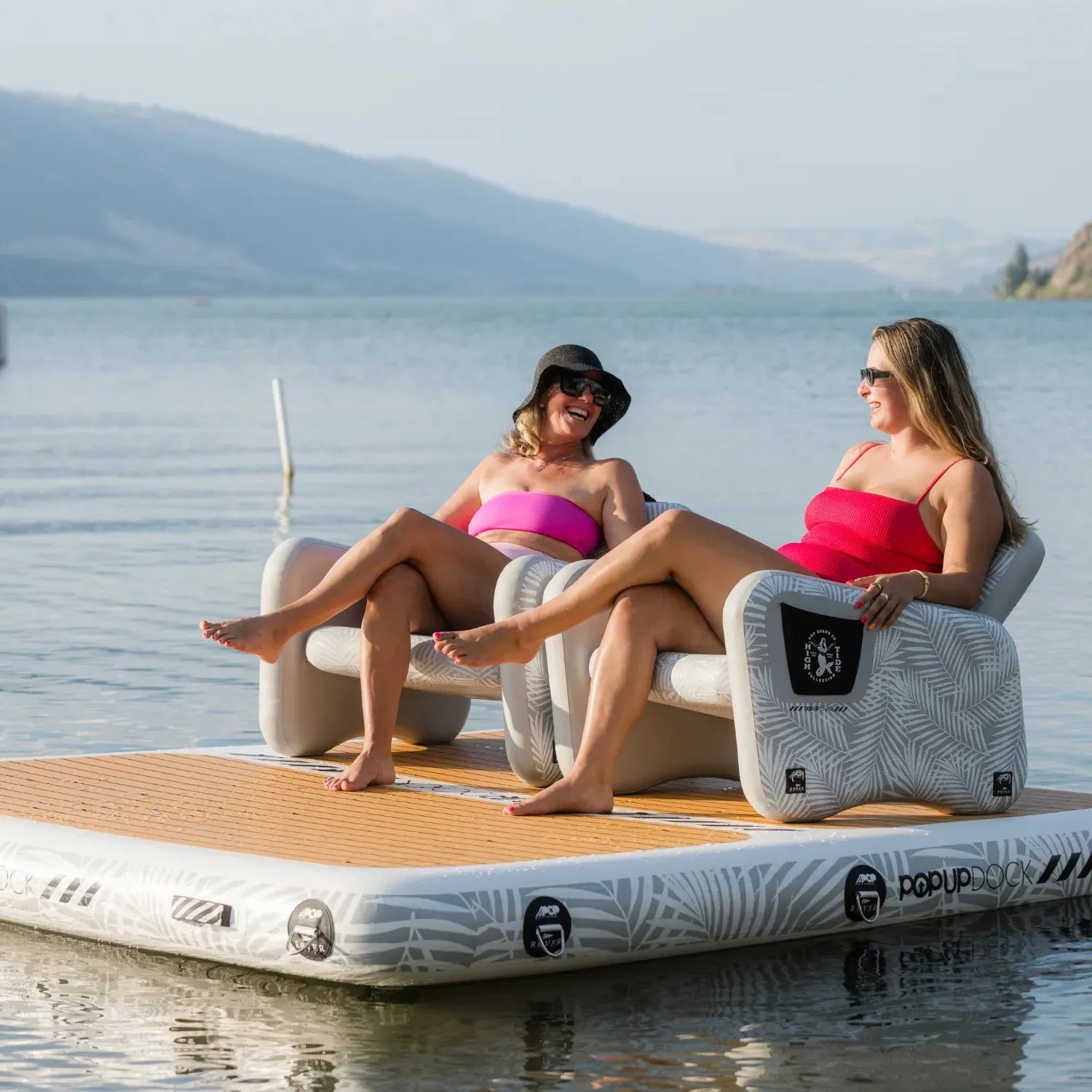 Two women in swimsuits lounging on inflatable chairs placed on a POPUP Dock floating platform, enjoying a sunny day on the water.