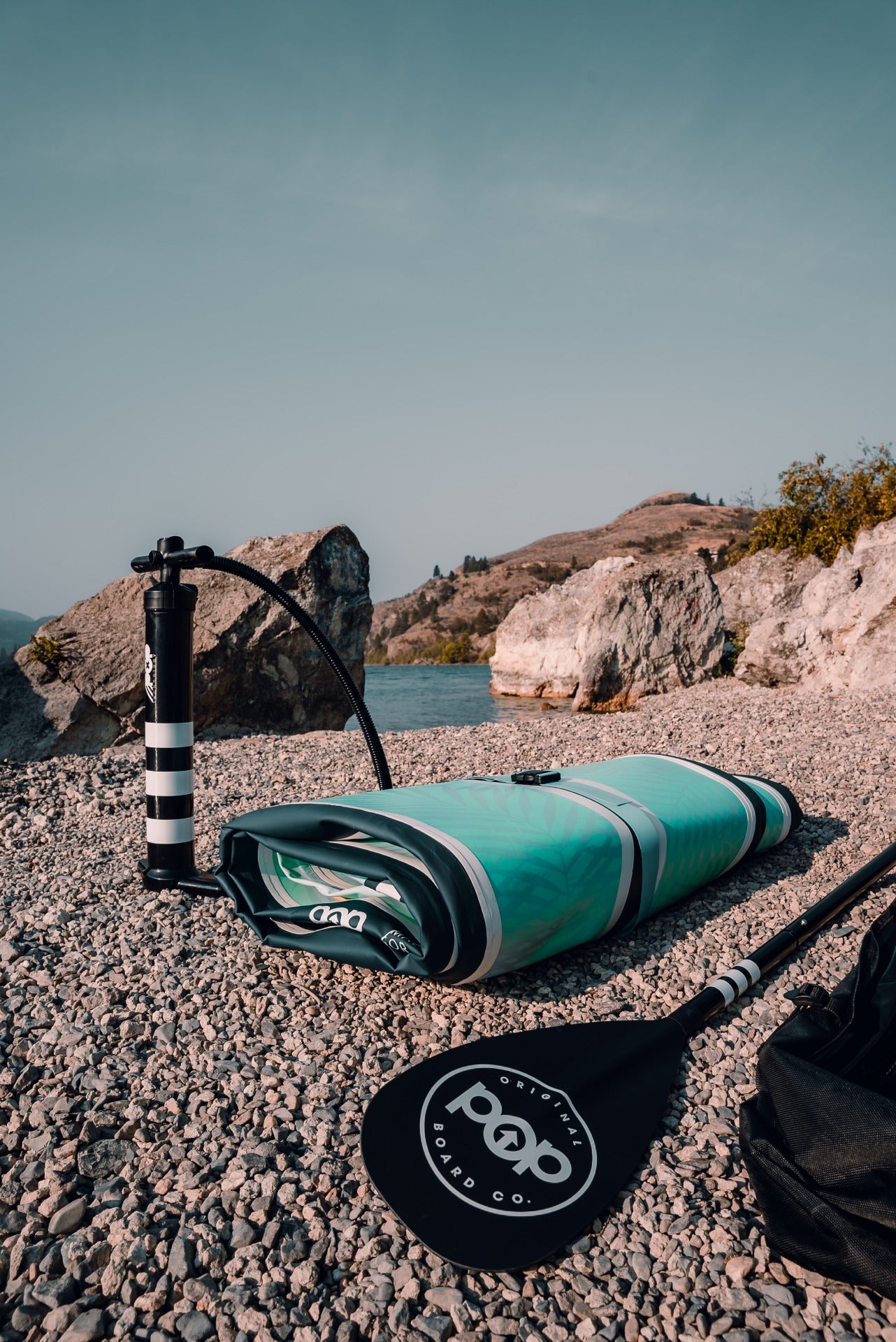 The 11'0" Yacht Hopper inflatable paddleboard deflated and rolled up on a rocky beach, displayed with a high-pressure hand pump and adjustable paddle, showing its compact portability.