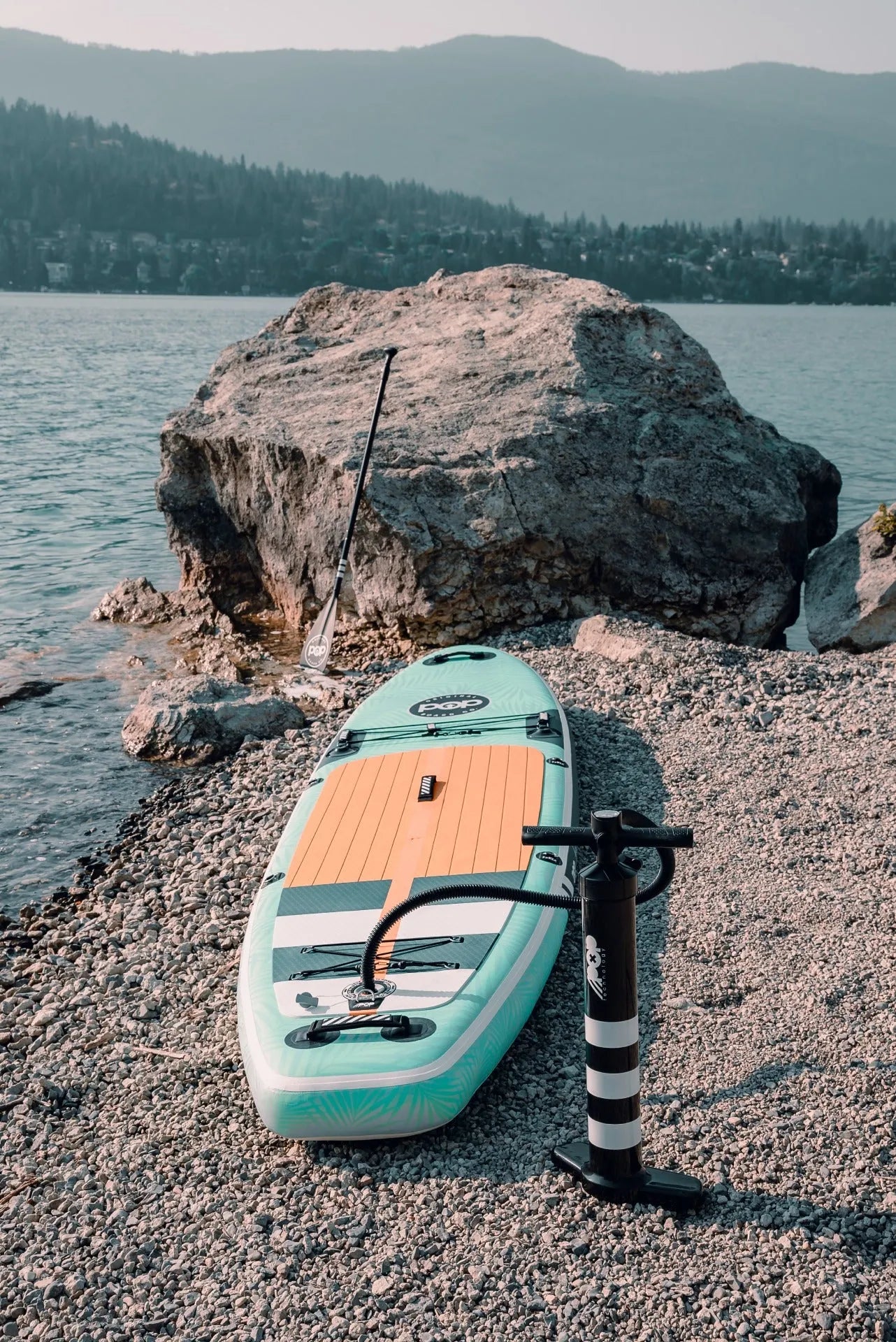The 11'0" Yacht Hopper inflatable paddleboard kit displayed on a rocky beach, including the inflated mint/teak board, a black high-pressure hand pump, and an adjustable paddle leaning against a rock.