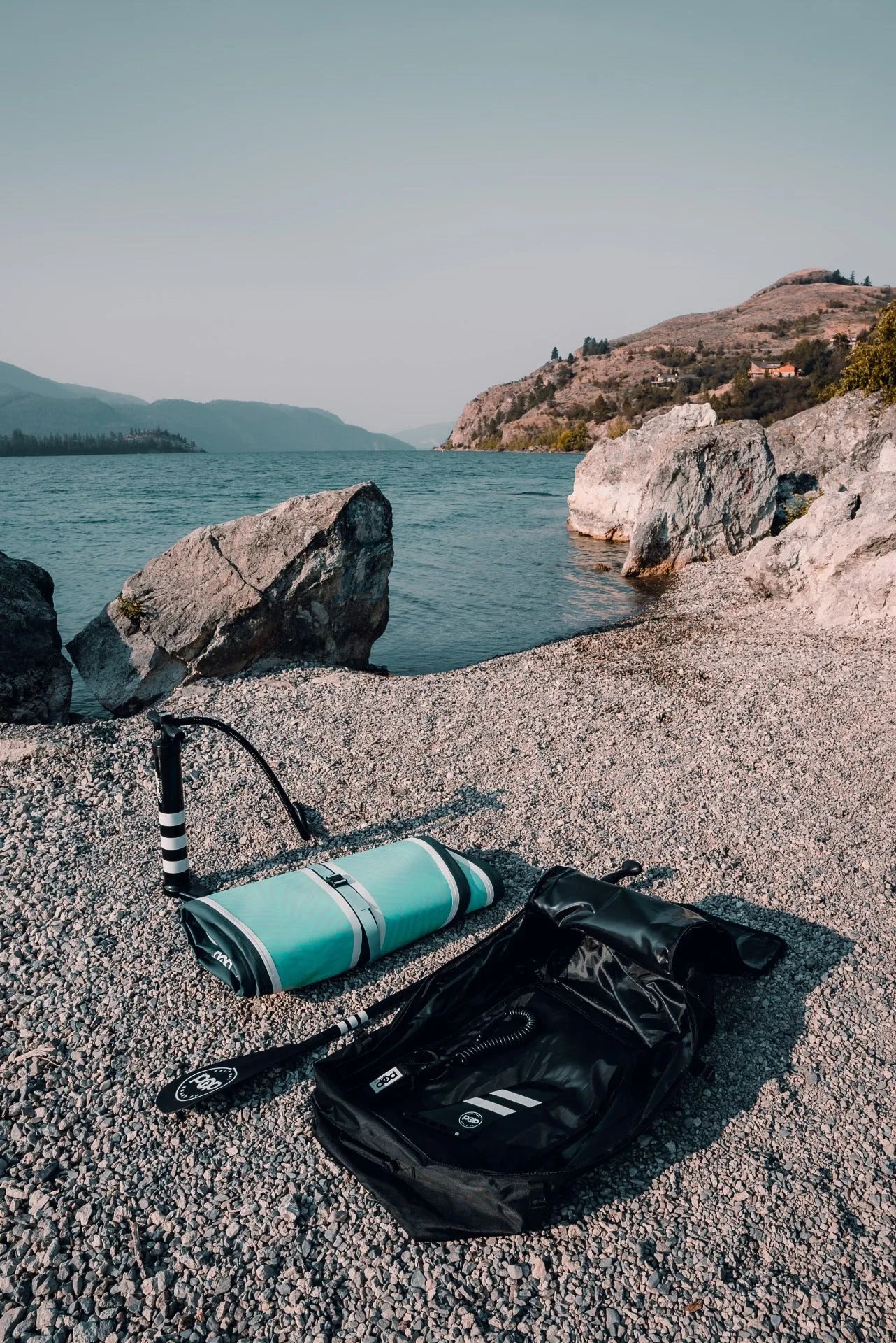 High-angle view of the deflated and rolled-up 11'0" Yacht Hopper inflatable paddleboard next to its open travel backpack, pump, and paddle on a rocky beach, demonstrating its portability.