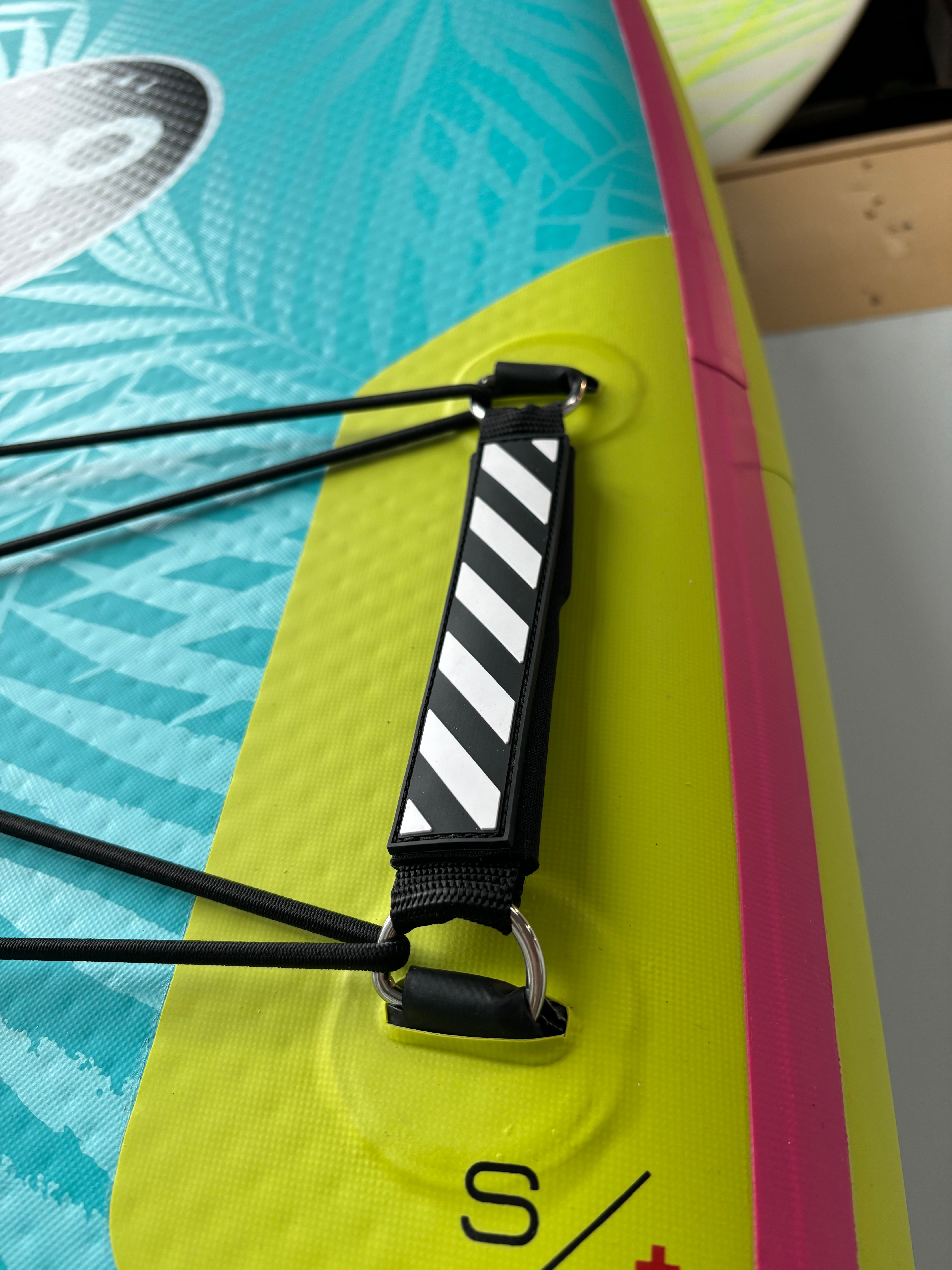 Close-up of the reinforced carry handle on the neon yellow and turquoise Yacht Hopper paddleboard, showing the black and white striped grip and integrated bungee cargo tie-downs.